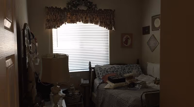 A dimly lit bedroom with a bed covered in patterned bedding and several pillows. There is a window with closed blinds and a floral valance above it. A bedside table with a lamp and various items is next to the bed. The walls have framed pictures hanging on them.