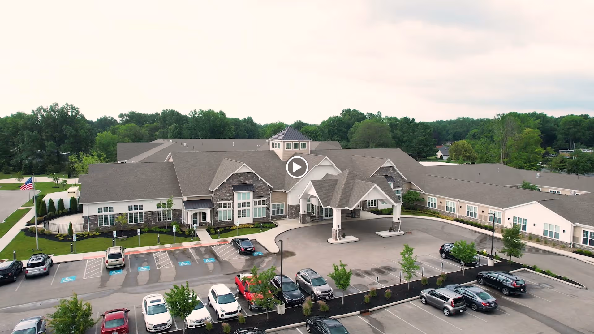 Aerial view of Vitalia Senior Residences Westlake showing a large single-story building with a covered entrance, surrounded by a parking lot with several cars and landscaped greenery. Trees and a partly cloudy sky are visible in the background.