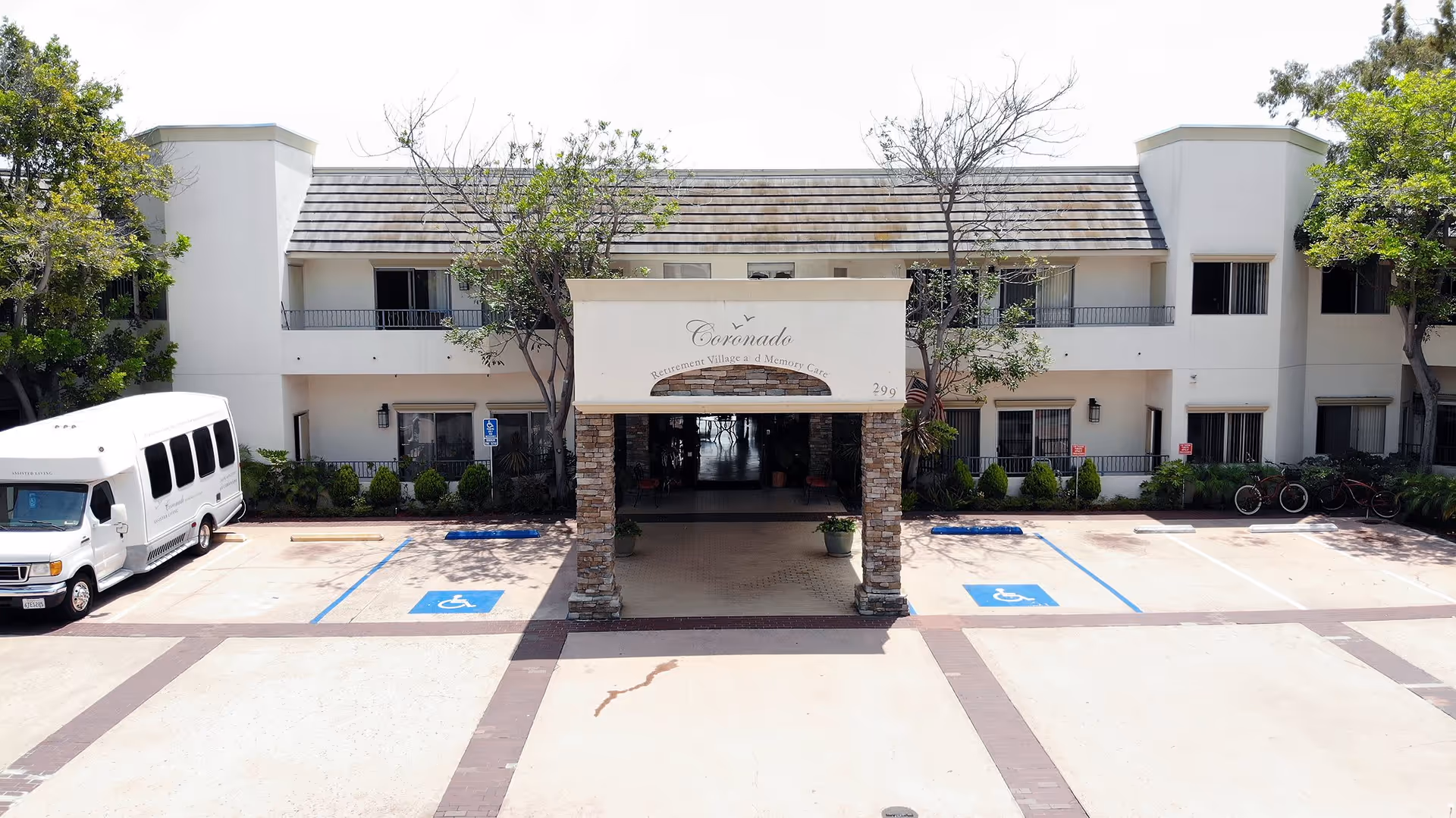 Front exterior view of Coronado Retirement Village building with a covered entrance supported by stone pillars, surrounded by trees and a parking lot with handicap parking spaces and a white shuttle bus parked on the left side.