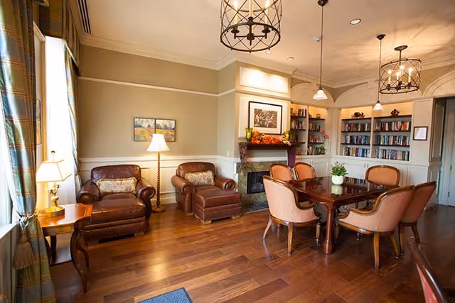 A cozy living room area with two brown leather armchairs and an ottoman, a wooden side table with a lamp, a wooden dining table with six upholstered chairs, built-in bookshelves filled with books, a fireplace decorated with autumn-themed items, and warm lighting from ceiling fixtures and lamps.