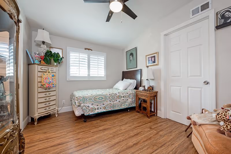 A cozy bedroom with a single bed covered in a patterned bedspread, a wooden nightstand with a lamp, a tall dresser with a plant and birthday card on top, a ceiling fan with light, wooden flooring, and framed pictures on the walls.