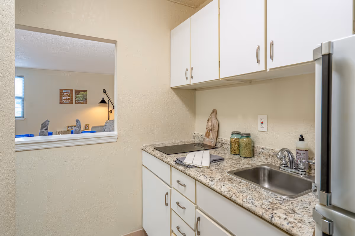 A small kitchen area with white cabinets, a granite countertop, a stainless steel sink, and a stovetop. There are two jars with pasta and grains on the counter, a soap dispenser, and a towel. A window-like opening in the wall shows a glimpse of a living room with a lamp, wall art, and a window.