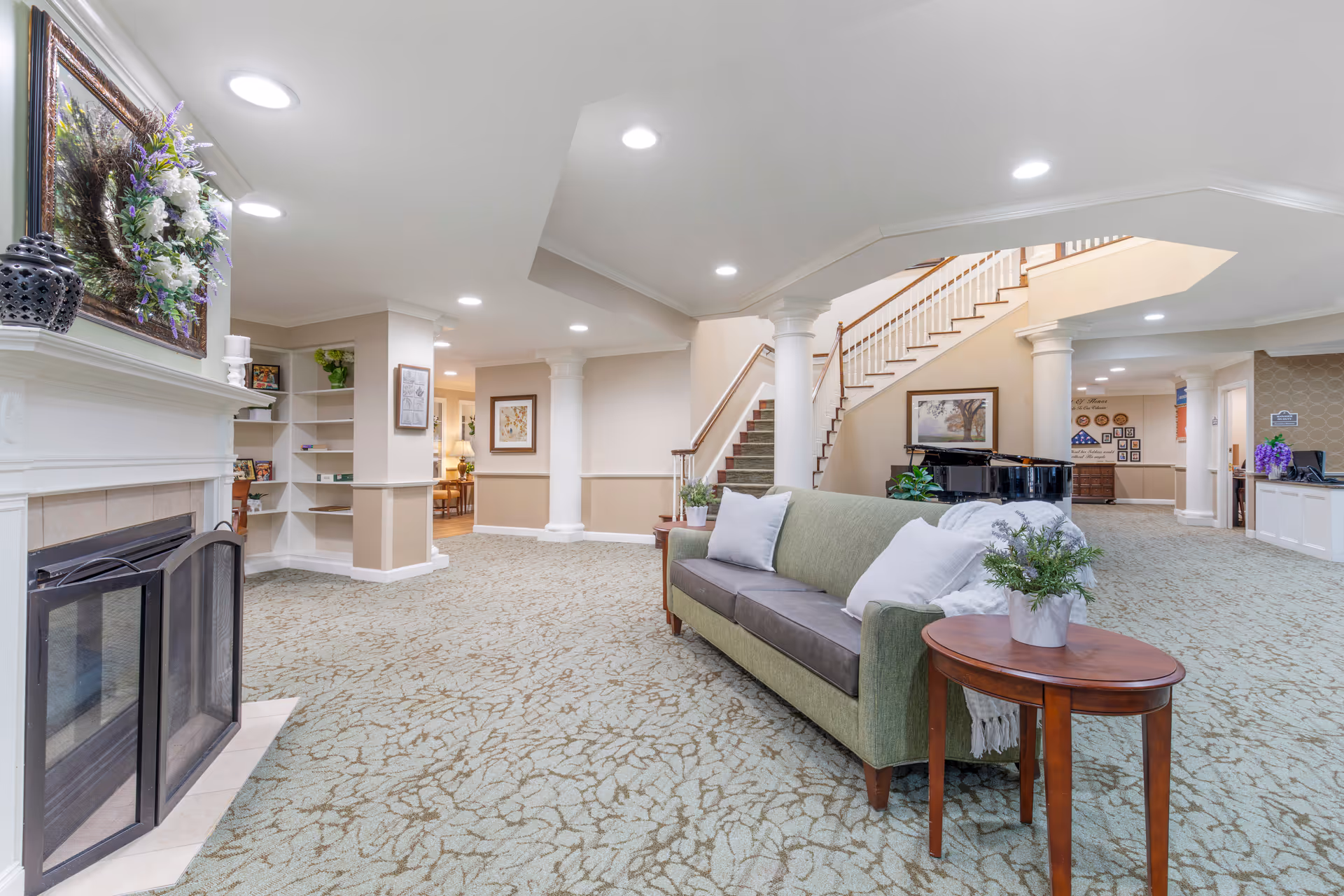 A spacious and well-lit living area in a senior living facility featuring a green sofa with white pillows and a throw blanket, a small wooden side table with a potted plant, a fireplace with a floral wreath above it, white columns, a staircase with wooden handrails, and a black grand piano in the background.