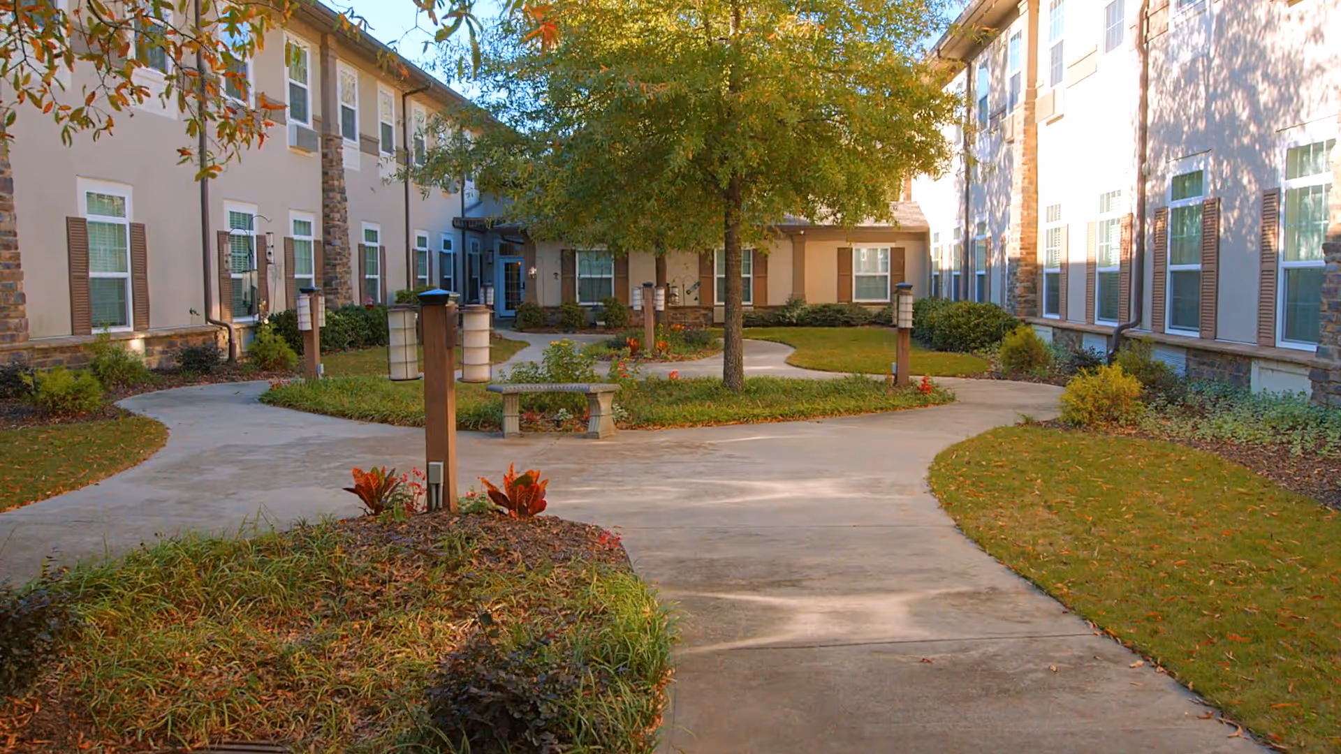 Landscaped courtyard with curved walkways, benches, lamp posts, and a central tree surrounded by a two-story residential building.