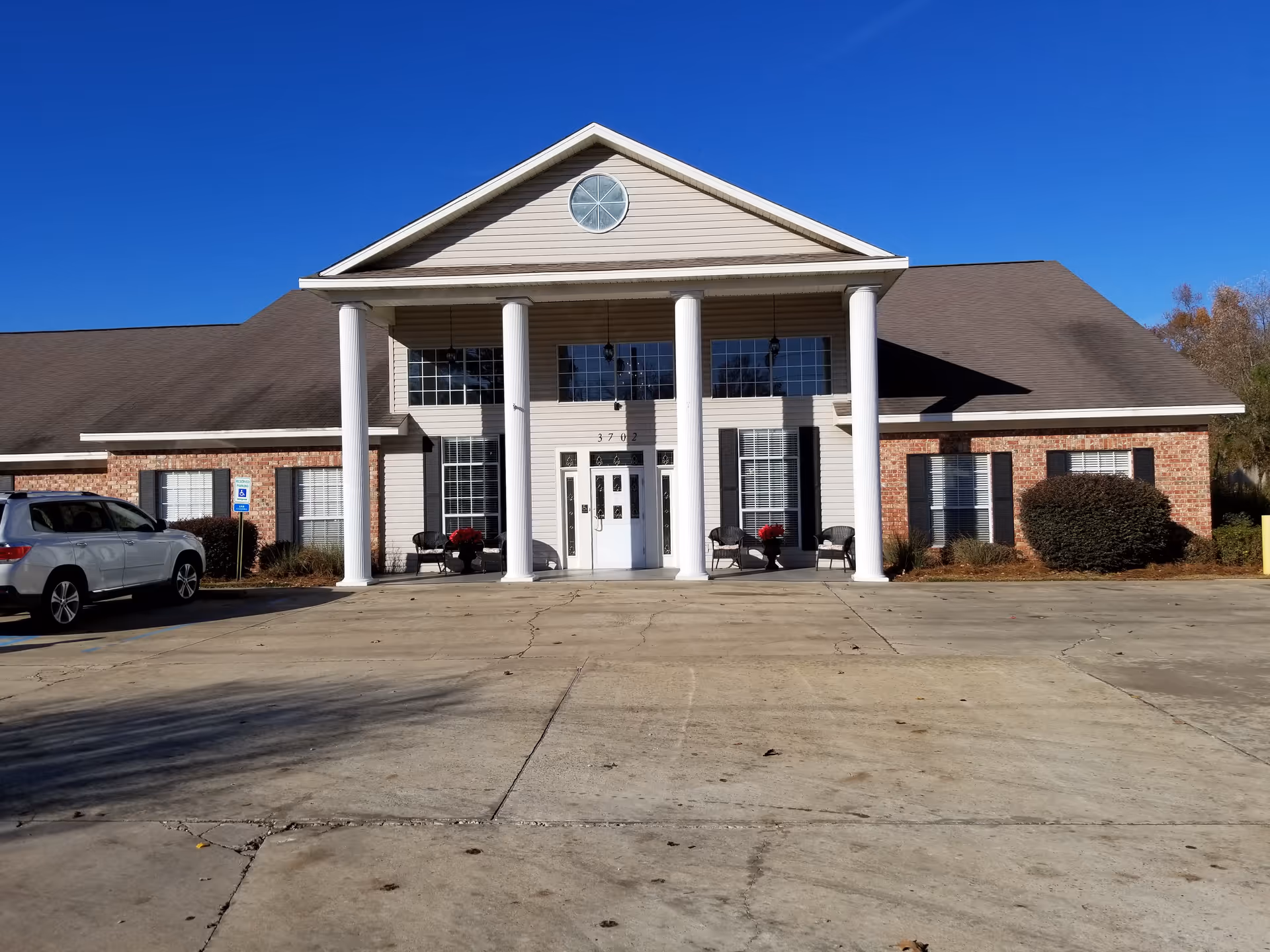 Front entrance of a single-story brick and siding building with white columns, seating, and a parking area under a clear blue sky.