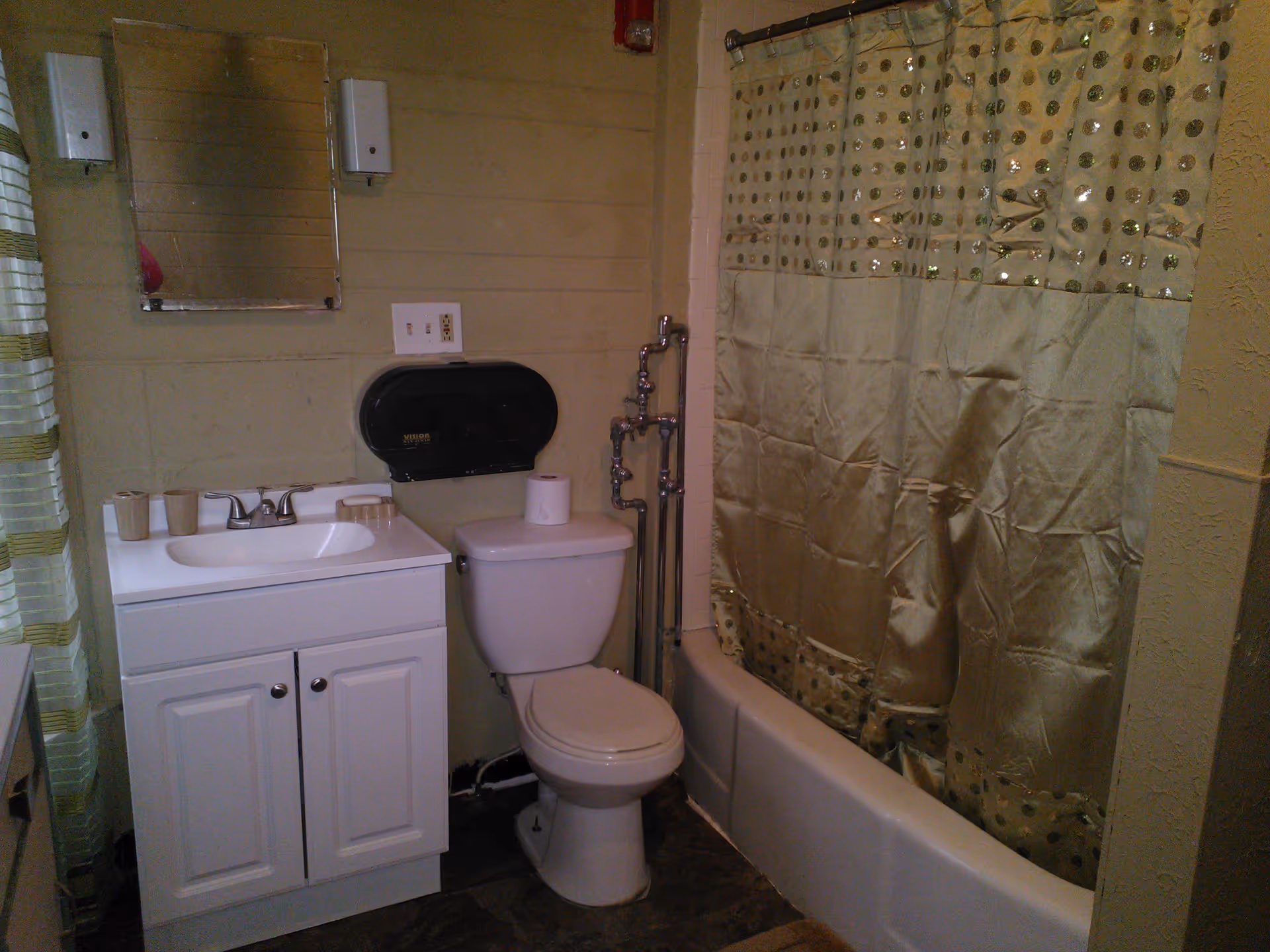 Small bathroom with a white vanity and sink, a toilet beneath a wall-mounted dispenser, and a bathtub with a patterned shower curtain.
