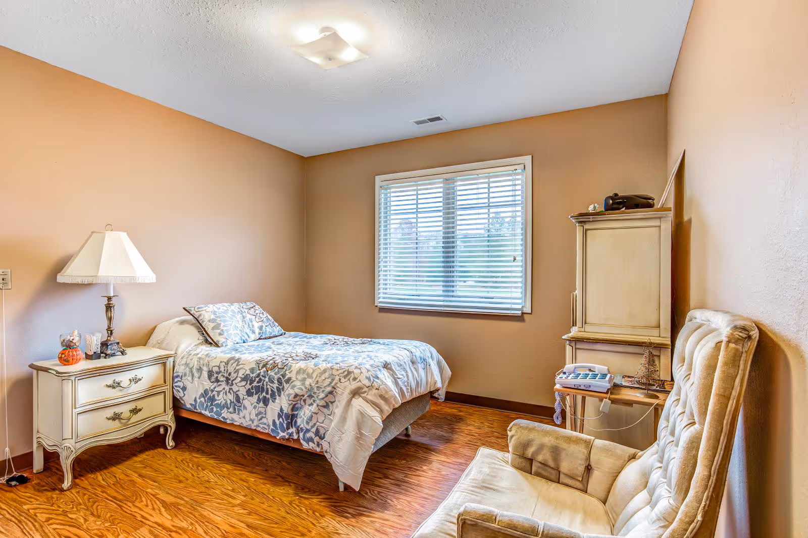 A cozy bedroom with a single bed covered in a floral patterned comforter, a nightstand with a lamp and decorative items, a window with blinds, a wooden cabinet with a telephone on top, and a cushioned armchair.