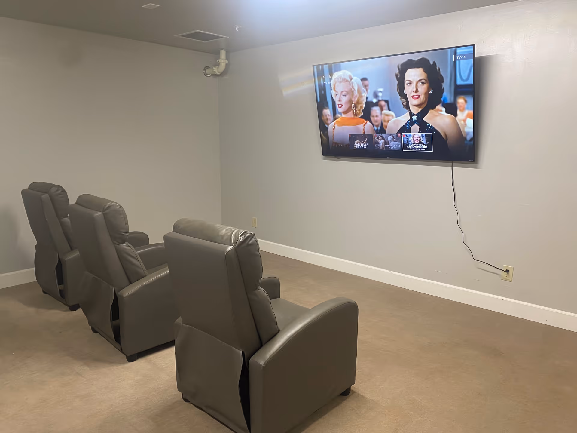 A small media room with four gray recliner chairs arranged in two rows facing a wall-mounted flat screen TV displaying a classic movie scene. The room has plain light-colored walls and a beige carpeted floor.