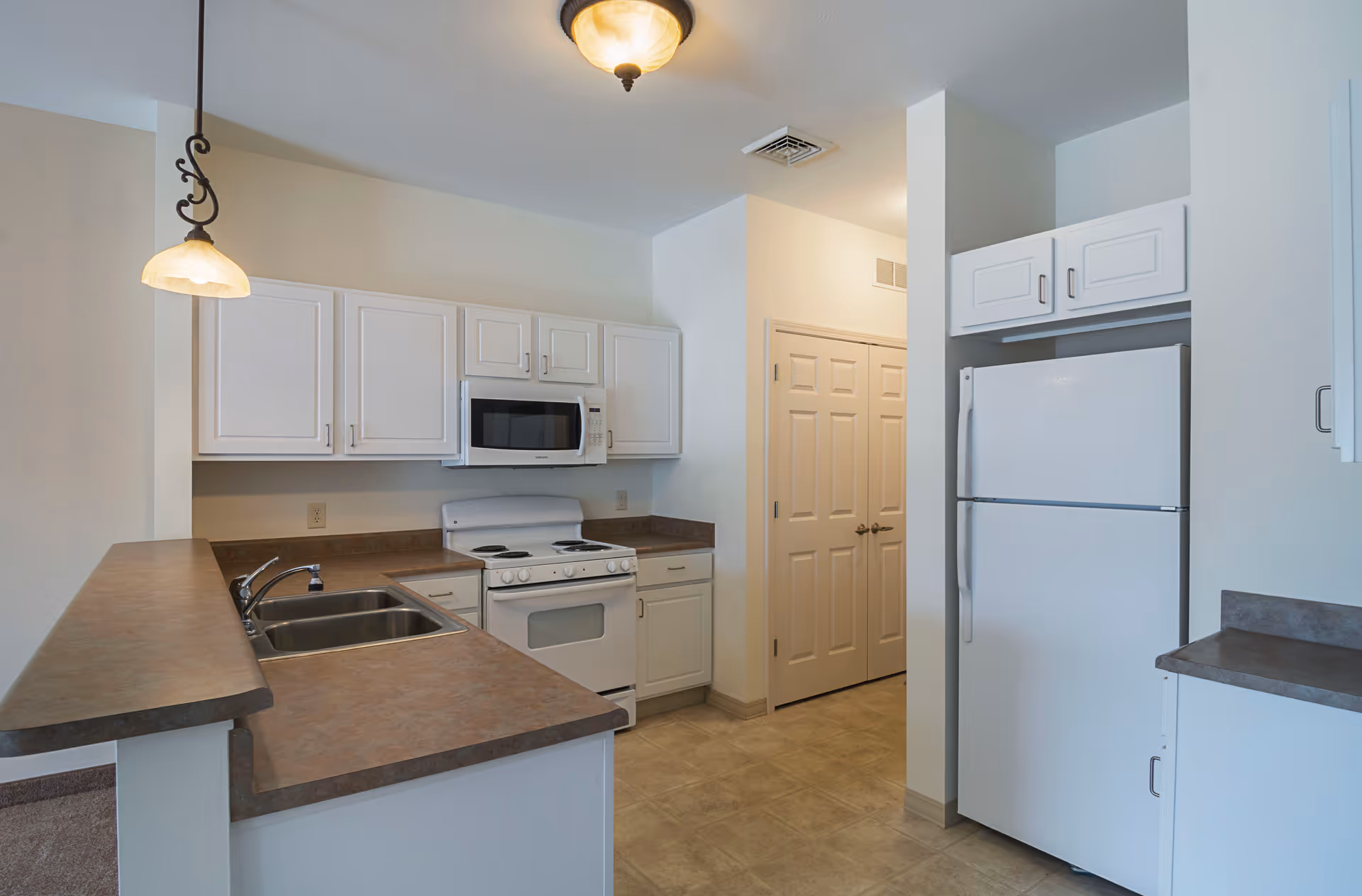 A clean and modern kitchen featuring white cabinets, a white refrigerator, a white stove with an oven, a microwave above the stove, a double sink, and brown countertops. The kitchen has a hanging light fixture and beige tiled flooring.