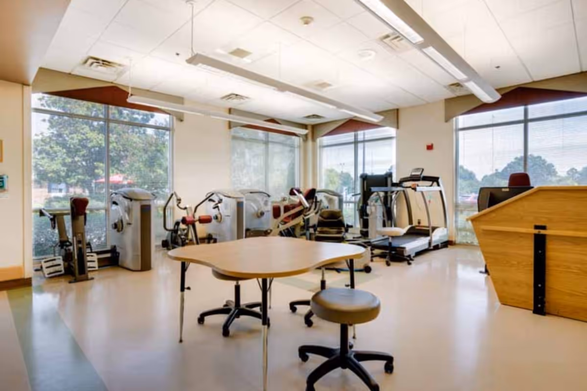 A bright exercise room with large windows showing outdoor greenery. The room contains various exercise machines including a treadmill, stationary bikes, and strength training equipment. In the center, there is a wooden table with two rolling stools. The floor is light-colored and the ceiling has fluorescent lighting.
