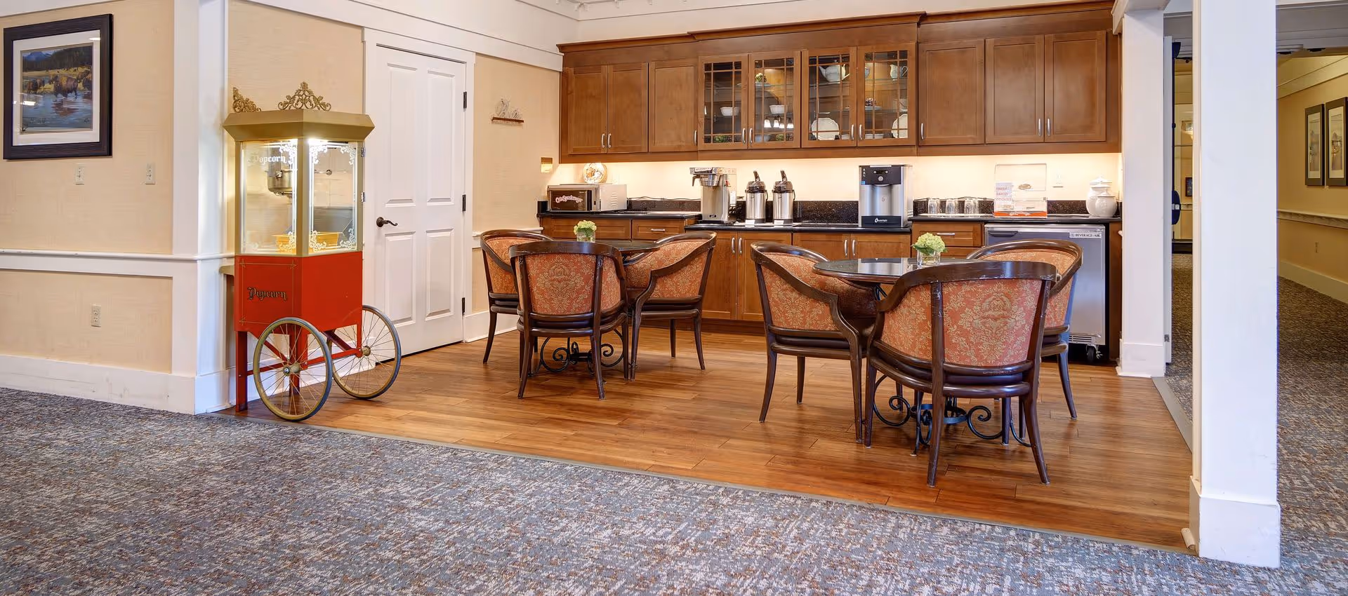 A cozy common area with wooden flooring and carpet, featuring two round tables each surrounded by four upholstered chairs with patterned fabric. Behind the tables is a wooden cabinet with glass doors displaying dishes, and a countertop with coffee dispensers, a microwave, and other kitchen appliances. To the left, there is a vintage-style popcorn machine on wheels. The walls are beige with white trim, and a framed picture hangs on the left wall.