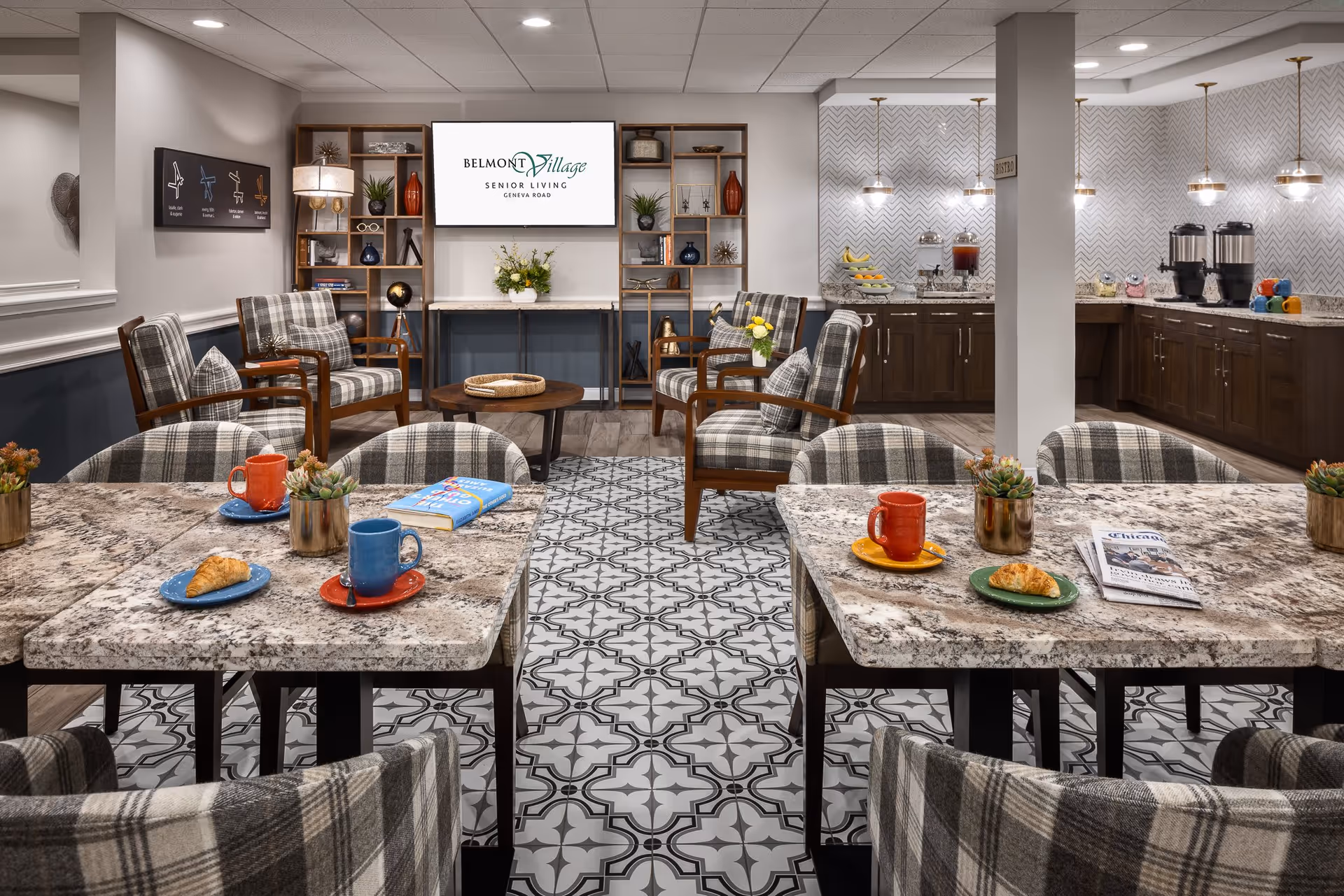 A cozy senior living common area with plaid upholstered chairs around granite tables set with colorful mugs, plates with croissants, and books. In the background, there are more armchairs arranged around a round wooden coffee table, a shelving unit with decorative items, and a large screen displaying 'Belmont Village Senior Living Geneva Road'. To the right, a counter area with coffee dispensers, fruit, and other refreshments is visible under pendant lights.