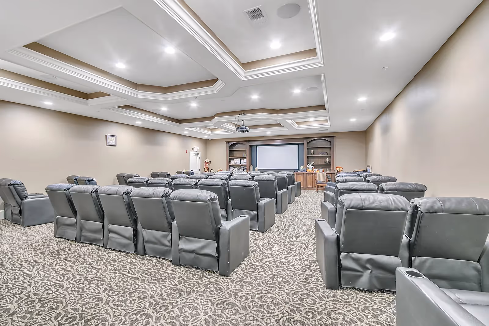 A large room with multiple rows of black leather recliner chairs facing a large projection screen at the front. The room has a patterned carpet, beige walls, and a coffered ceiling with recessed lighting. There are built-in shelves and cabinets on either side of the screen.