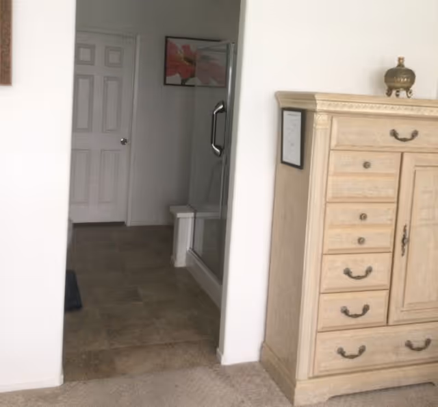 View of a hallway leading to a bathroom with a glass shower door and a closed white door. To the right, there is a light wood dresser with multiple drawers and decorative handles, topped with a small ornamental item. The floor transitions from carpet to tile near the bathroom entrance.