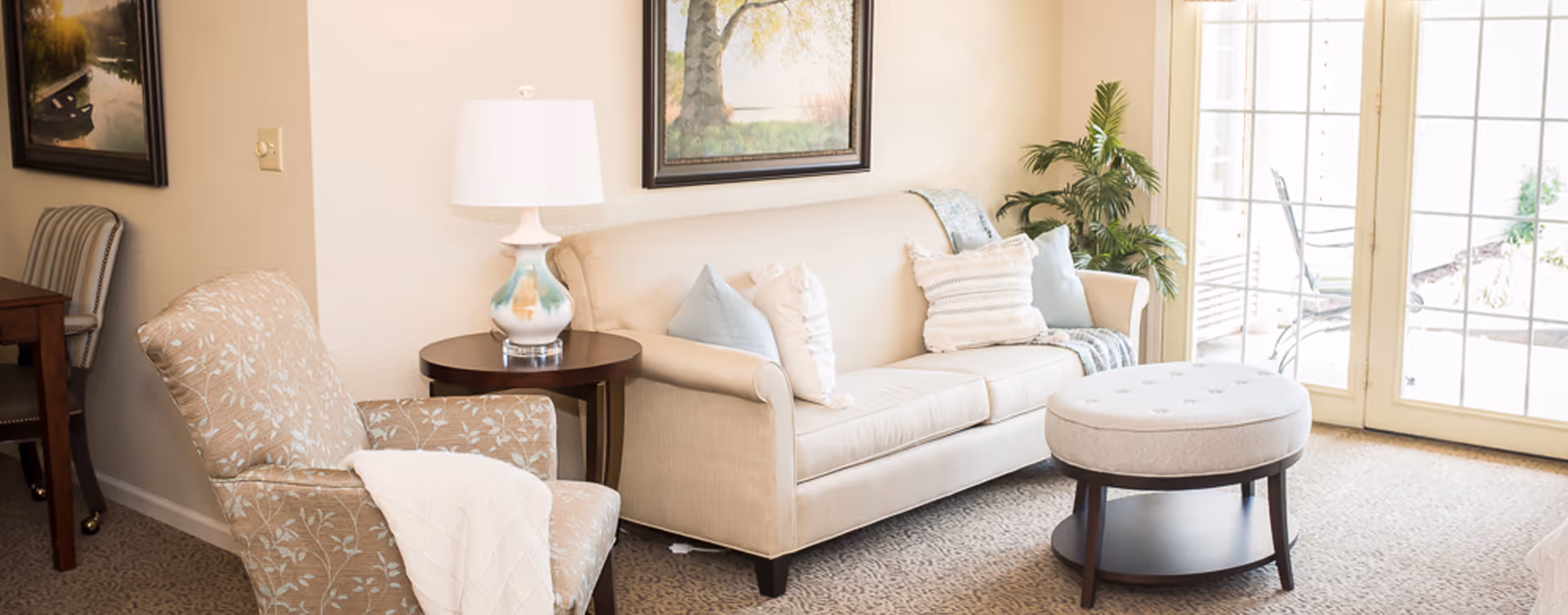A cozy living room with a beige sofa adorned with multiple pillows and a throw blanket, a patterned armchair with a white blanket draped over it, a round ottoman, a side table with a decorative lamp, a potted plant, and large glass doors letting in natural light.