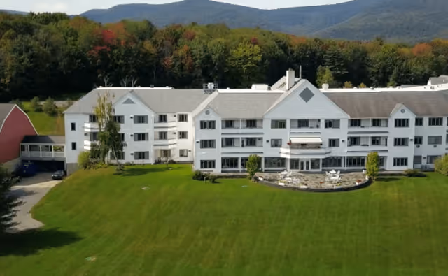 A large white multi-story building with numerous windows, surrounded by a well-maintained green lawn and trees in the background, with hills visible in the distance under a clear sky.