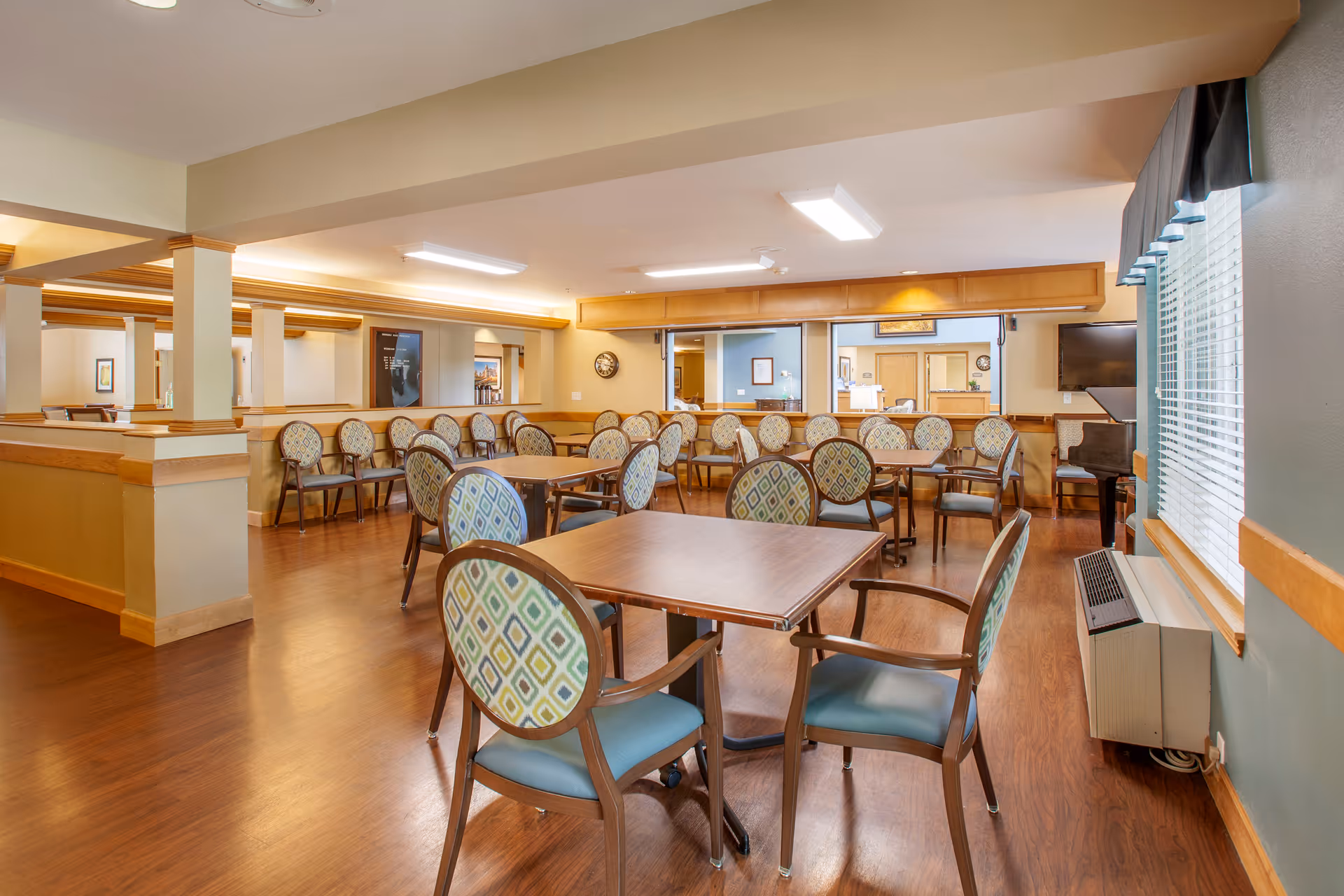 A spacious dining room with multiple square wooden tables and cushioned chairs featuring patterned backs and blue seats. The room has wood flooring, beige walls with wooden trim, and large windows with blinds on the right side. There is a piano in the back corner and a clock on the wall. The ceiling has recessed lighting and the room appears clean and well-lit.