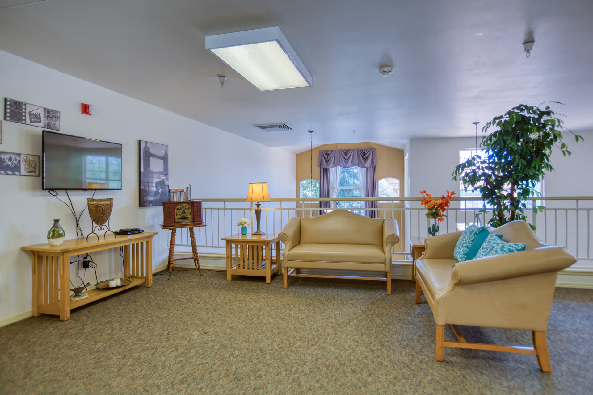 A cozy living room area with two beige sofas, a wooden side table with a lamp, a wooden TV stand with decorative items, a wall-mounted flat screen TV, framed pictures on the wall, and a large potted plant near a window with purple curtains.