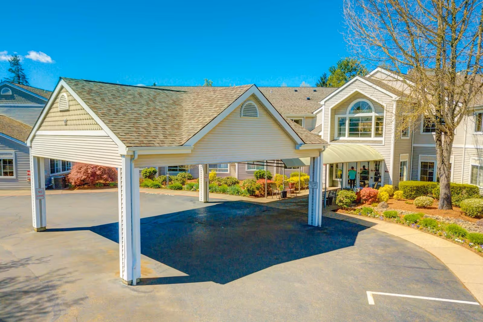 Covered porte-cochere at the front entrance of a multi-story senior living building with landscaping and a paved driveway.