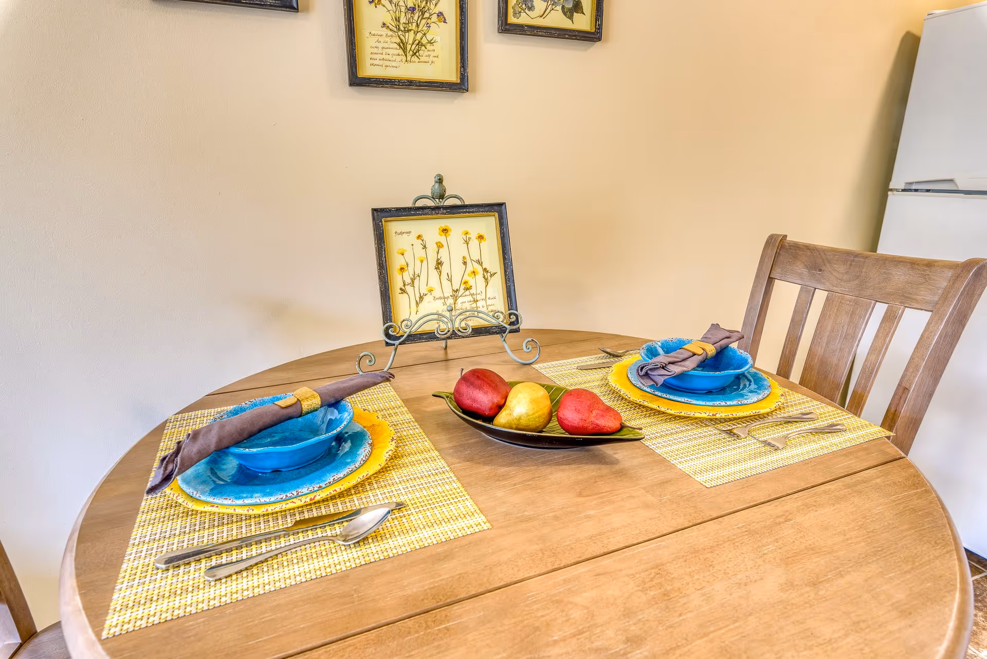 A wooden round dining table set for two with yellow and blue plates, blue bowls, and brown napkins with yellow rings. A decorative bowl with two red pears and one yellow pear is in the center of the table. Behind the table, there are framed botanical prints on a beige wall and a white refrigerator is visible to the right.