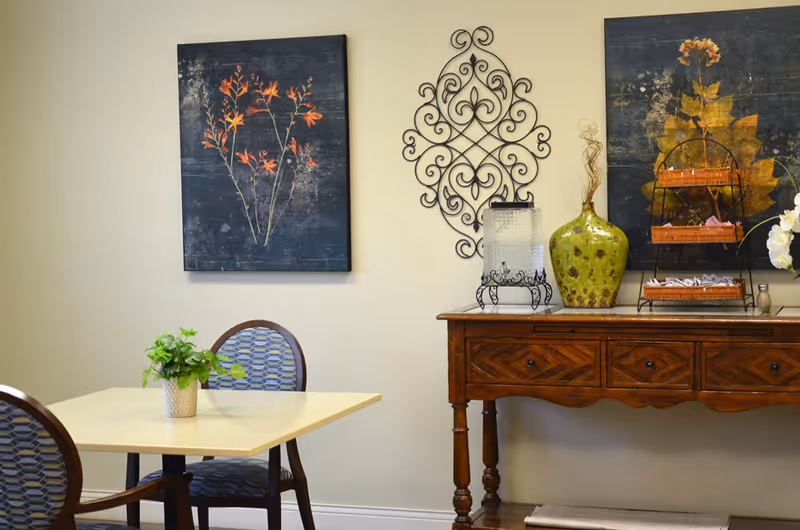 A small dining area with a square table and two chairs with patterned upholstery. On the table is a small potted plant. Against the wall is a wooden console table with three drawers, decorated with a large green vase, a water dispenser, a tiered basket with packets, and a small flower arrangement. The wall features two framed floral artworks and a decorative wrought iron piece.