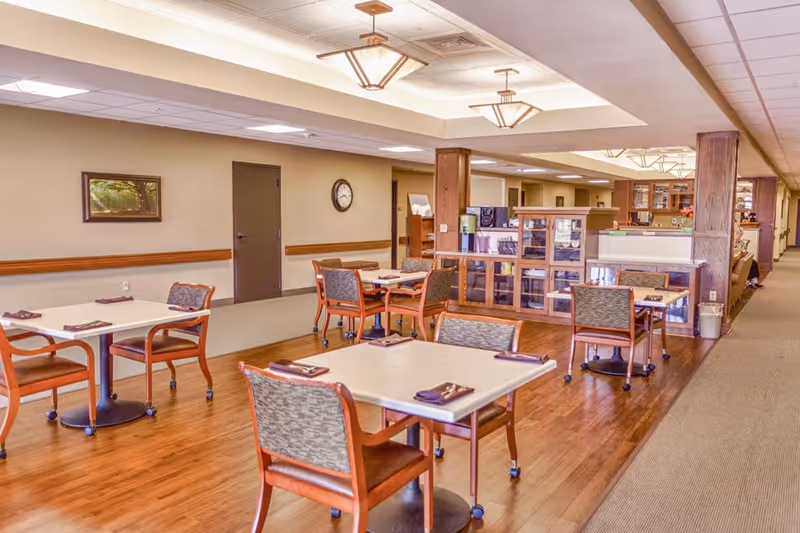 A dining area in a senior living facility with several square tables and chairs arranged neatly. Each table has a folded napkin and silverware. The room has wooden flooring, beige walls, a clock on the wall, and ceiling lights. There is a wooden partition with glass panels separating part of the space.