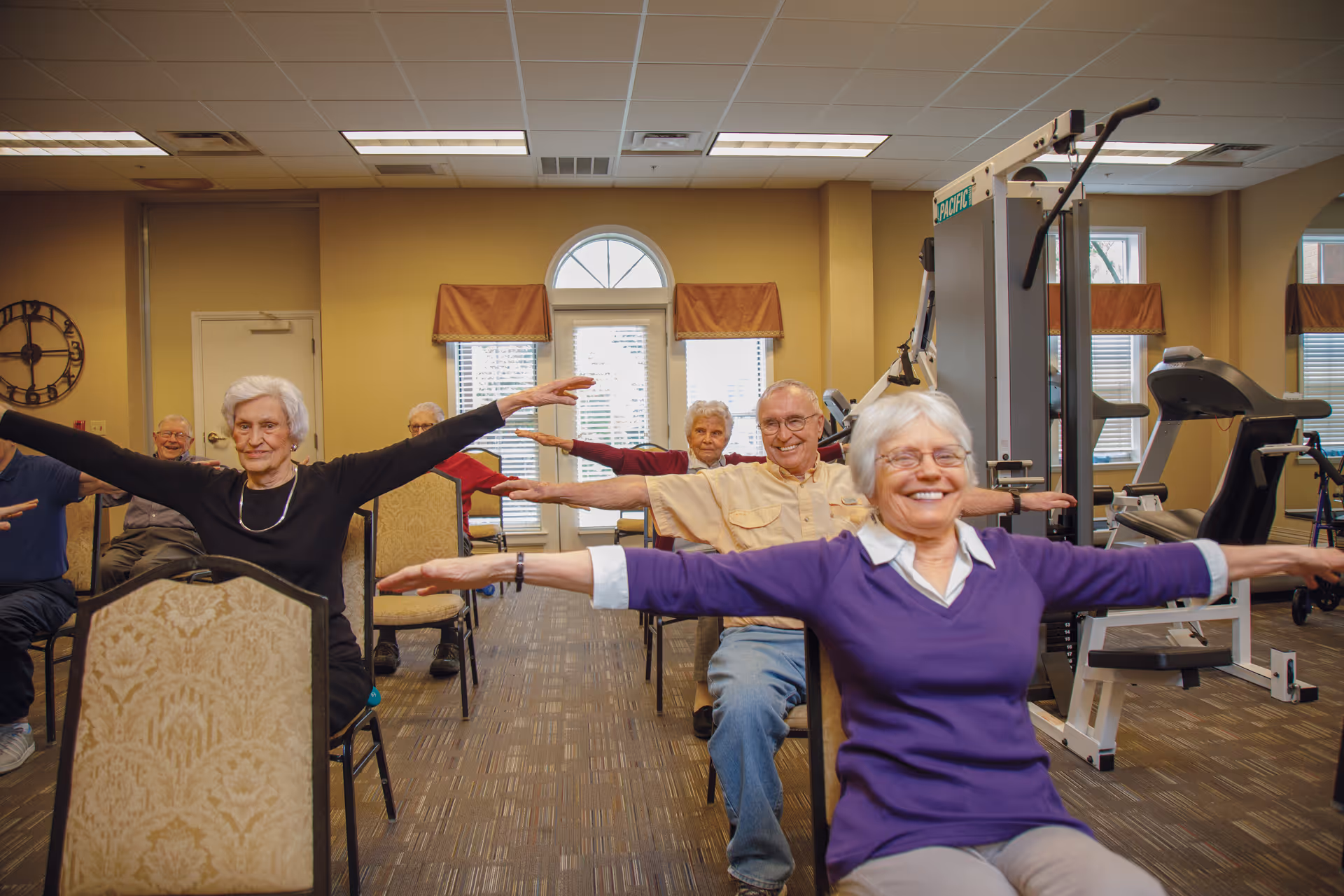 A group of elderly people sitting on chairs in a fitness room, participating in a seated exercise with their arms extended to the sides. The room has exercise equipment, windows with blinds and valances, and a clock on the wall.