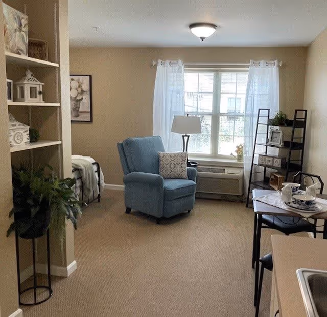 A cozy living space with a blue armchair and a decorative pillow facing a large window with sheer white curtains. To the right, there is a small dining table set with plates and cups, and a black shelving unit with decorative items. On the left, there is a partial view of a bed and a built-in shelving unit with various decorative pieces. The room has beige walls and carpeted flooring.