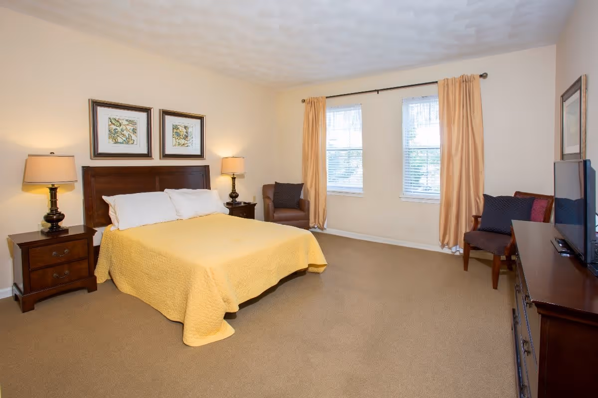 A bedroom with a bed covered in a yellow quilt, two white pillows, and a wooden headboard. There are two nightstands with lamps on either side of the bed. The room has beige walls, two windows with yellow curtains, a brown armchair, a wooden dresser with a flat-screen TV on top, and a chair with cushions.