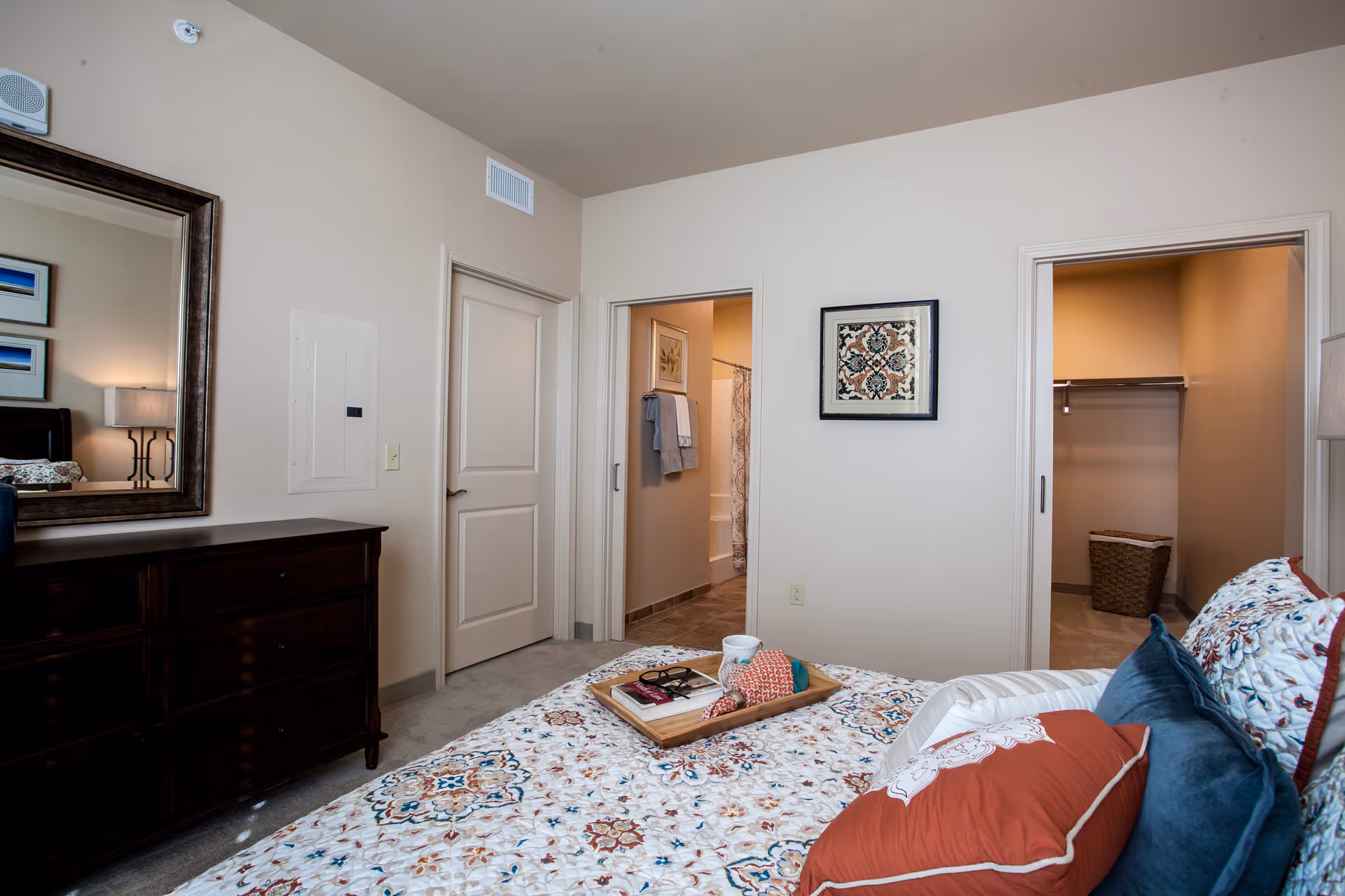 Well-appointed bedroom with patterned bedding, a dark dresser and open doors leading to a bathroom and a closet.