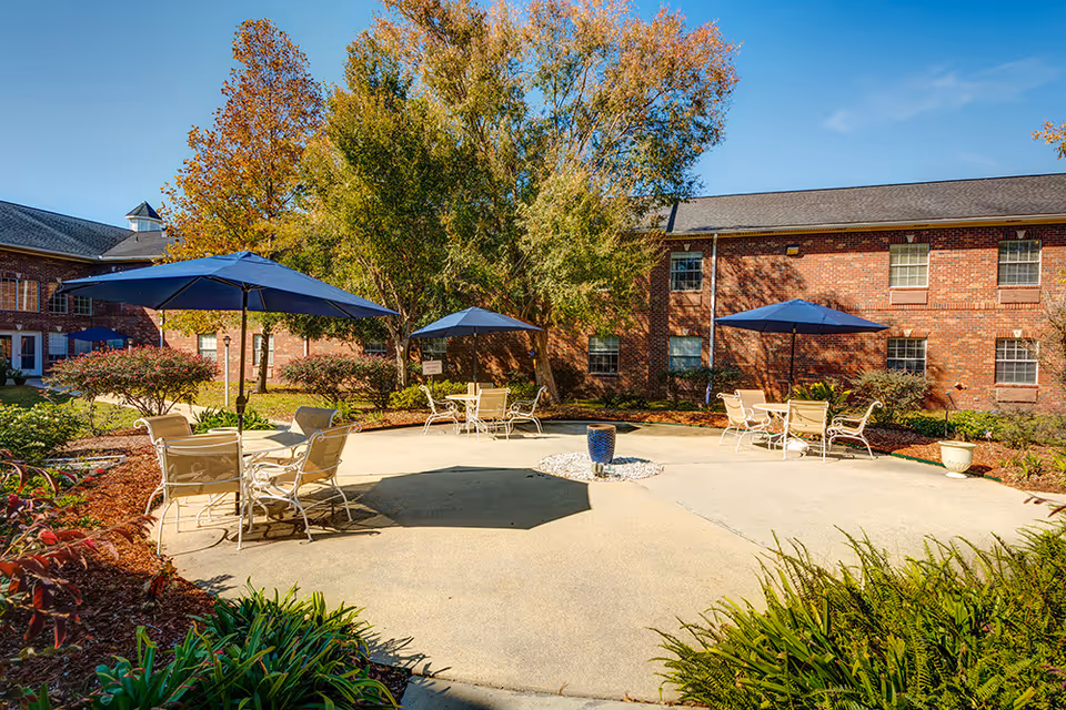 Sunny courtyard with patio tables and blue umbrellas in front of a two-story brick building.