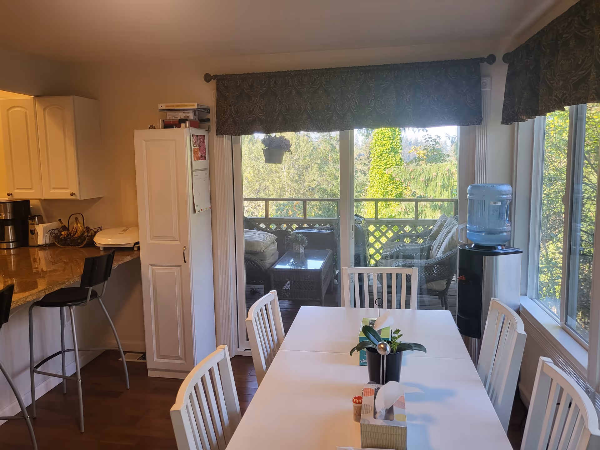 A bright dining area with a white table surrounded by white chairs. On the table are tissue boxes and a small potted plant. To the left is a kitchen counter with two black bar stools, a coffee maker, and a fruit basket. Large sliding glass doors lead to a balcony with wicker furniture and greenery outside. A water dispenser is positioned near a window on the right side.