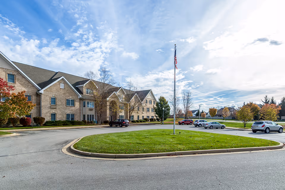 Exterior view of West Lafayette Assisted Living facility showing a large brick building with multiple windows and a peaked roof. In front of the building is a circular driveway with a grassy island in the center featuring a flagpole with an American flag. Several cars are parked along the driveway and trees with autumn foliage surround the area under a partly cloudy sky.
