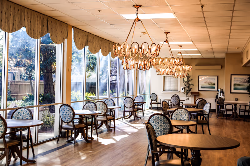 Sunlit dining room with round tables and patterned chairs, large windows along one wall, and decorative chandeliers.