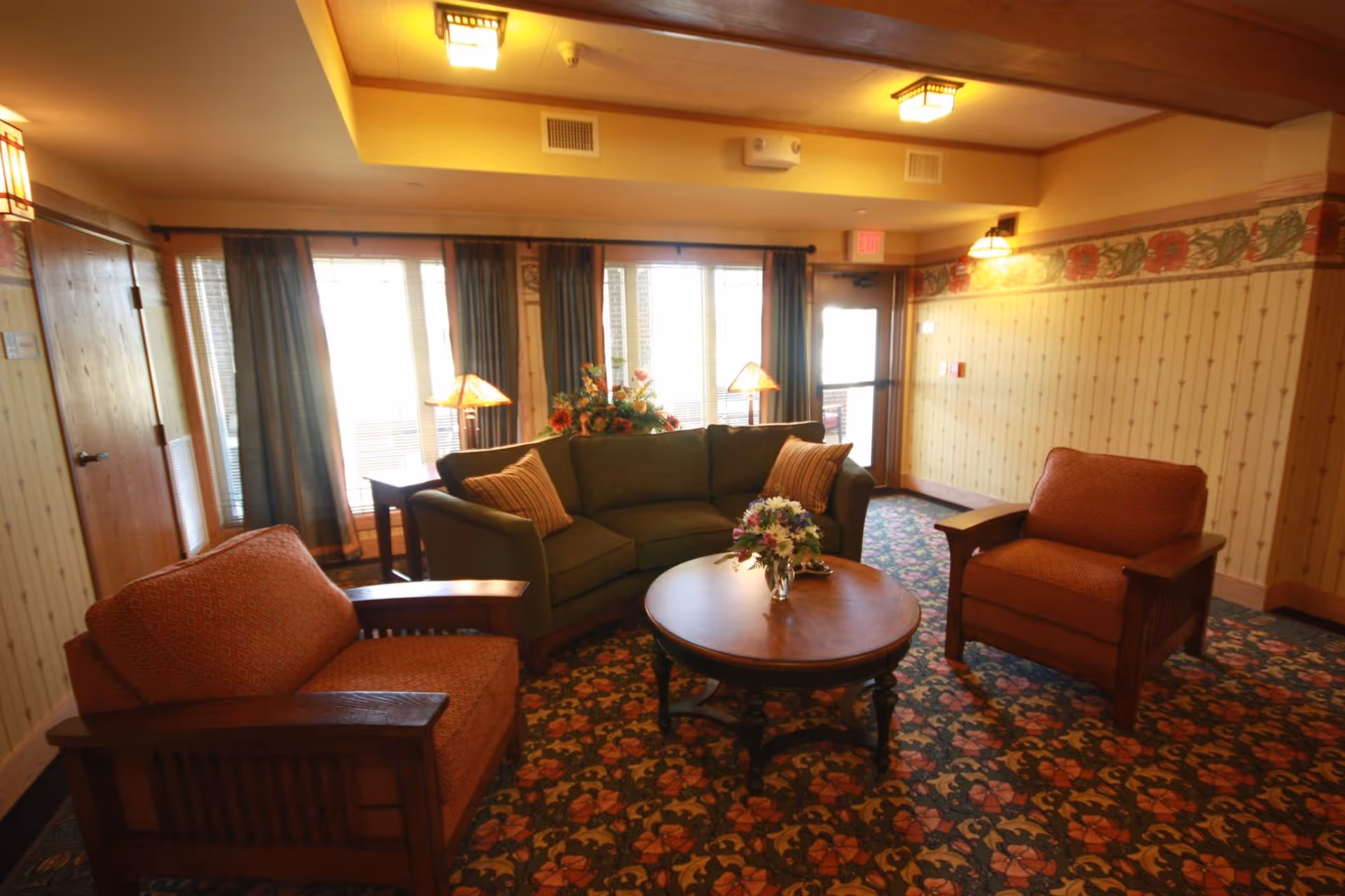 Cozy sitting area with a sofa, two armchairs and a round coffee table in a carpeted lounge with floral arrangements and windows in the background.