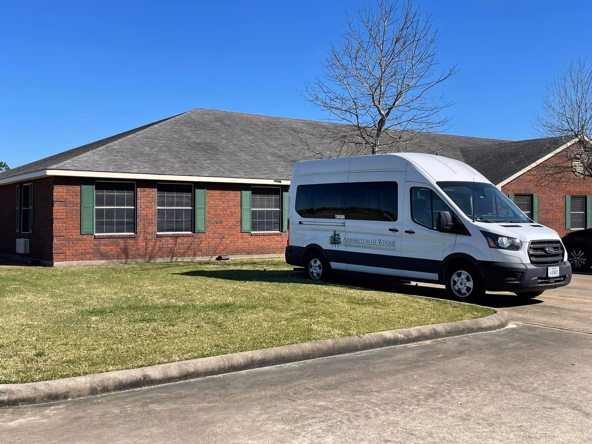 Exterior view of a single-story brick building with green window shutters under a clear blue sky. A white van with the logo and text 'Arboretum of Winnie' is parked on the driveway in front of the building. There are leafless trees near the building and a grassy area beside the driveway.