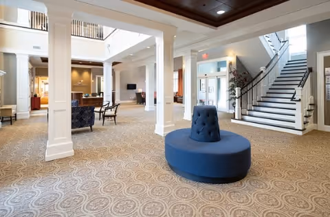 Spacious and well-lit assisted living facility lobby with patterned carpet, white columns, a circular blue cushioned bench in the center, seating areas with chairs and tables, a staircase with black railing, and large windows allowing natural light.