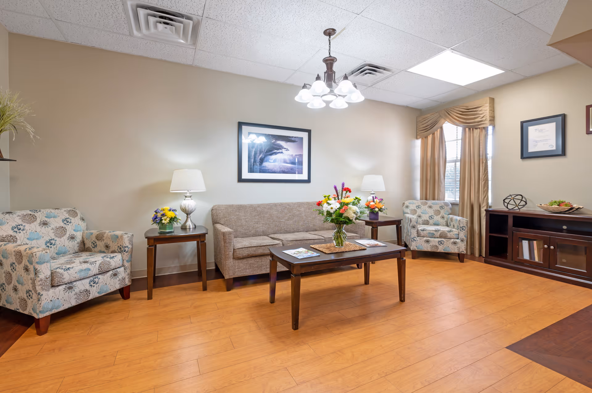 A cozy living room area with a beige sofa, two floral patterned armchairs, a wooden coffee table with a vase of colorful flowers, two side tables with lamps and flower arrangements, a framed picture on the wall, a window with beige curtains, and a wooden cabinet with books and decorative items.