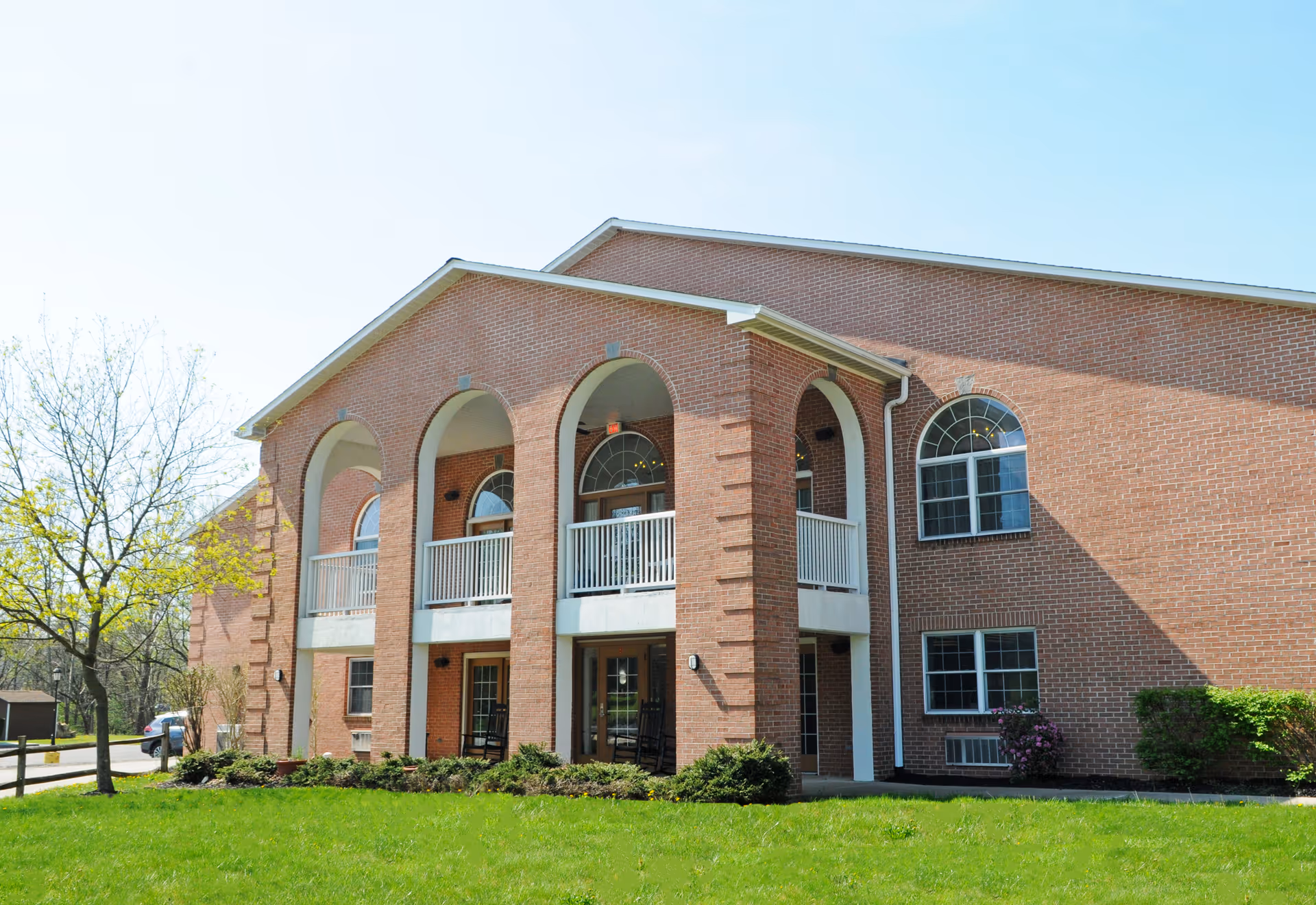 Exterior view of a two-story brick building with arched windows and a covered balcony supported by large arches. The building is surrounded by green grass, bushes, and a tree with budding leaves under a clear blue sky.