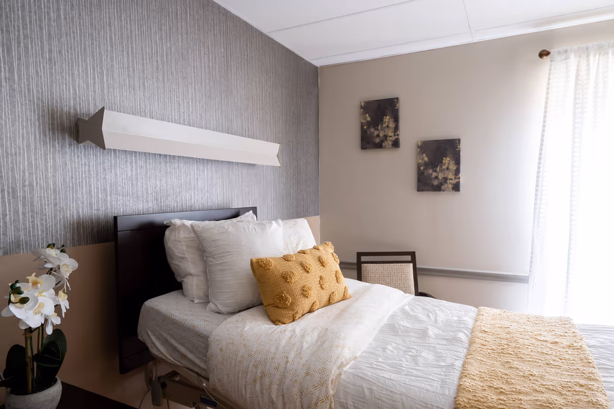 A neatly made bedroom in a senior living facility with a bed, decorative pillow, chair, orchid, wall art, and sheer curtains.