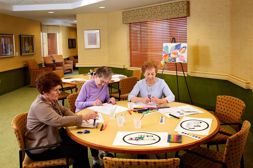 Three elderly women sitting around a round table in a well-lit room, engaged in coloring and painting activities with various art supplies spread on the table. The room has green and beige walls, patterned chairs, framed artwork on the walls, and a window with wooden blinds and a colorful painting on an easel nearby.