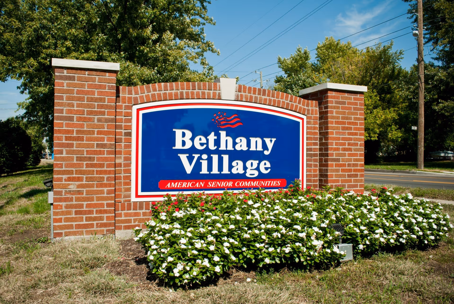A brick sign for Bethany Village, an American senior community, surrounded by green bushes with white flowers, with a clear blue sky and trees in the background.