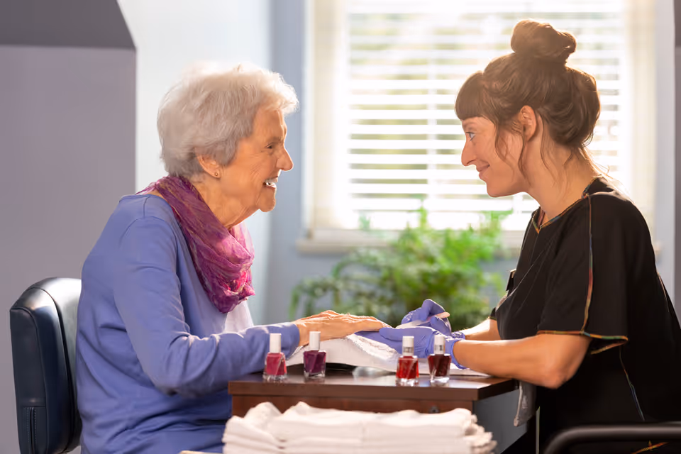 An elderly woman with white hair and a purple scarf is smiling while getting a manicure from a younger woman wearing gloves. They are seated at a table with several bottles of nail polish and towels, with a window and green plants in the background.