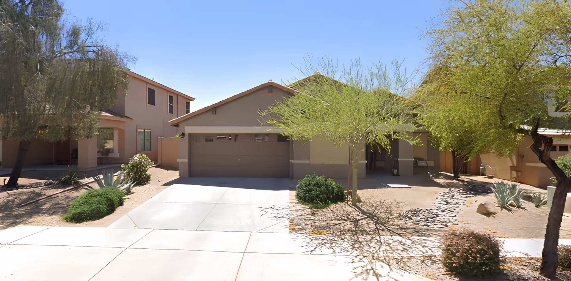 Front exterior view of a single-story residential building with a two-car garage, surrounded by desert landscaping including small bushes, trees, and rocks under a clear blue sky.