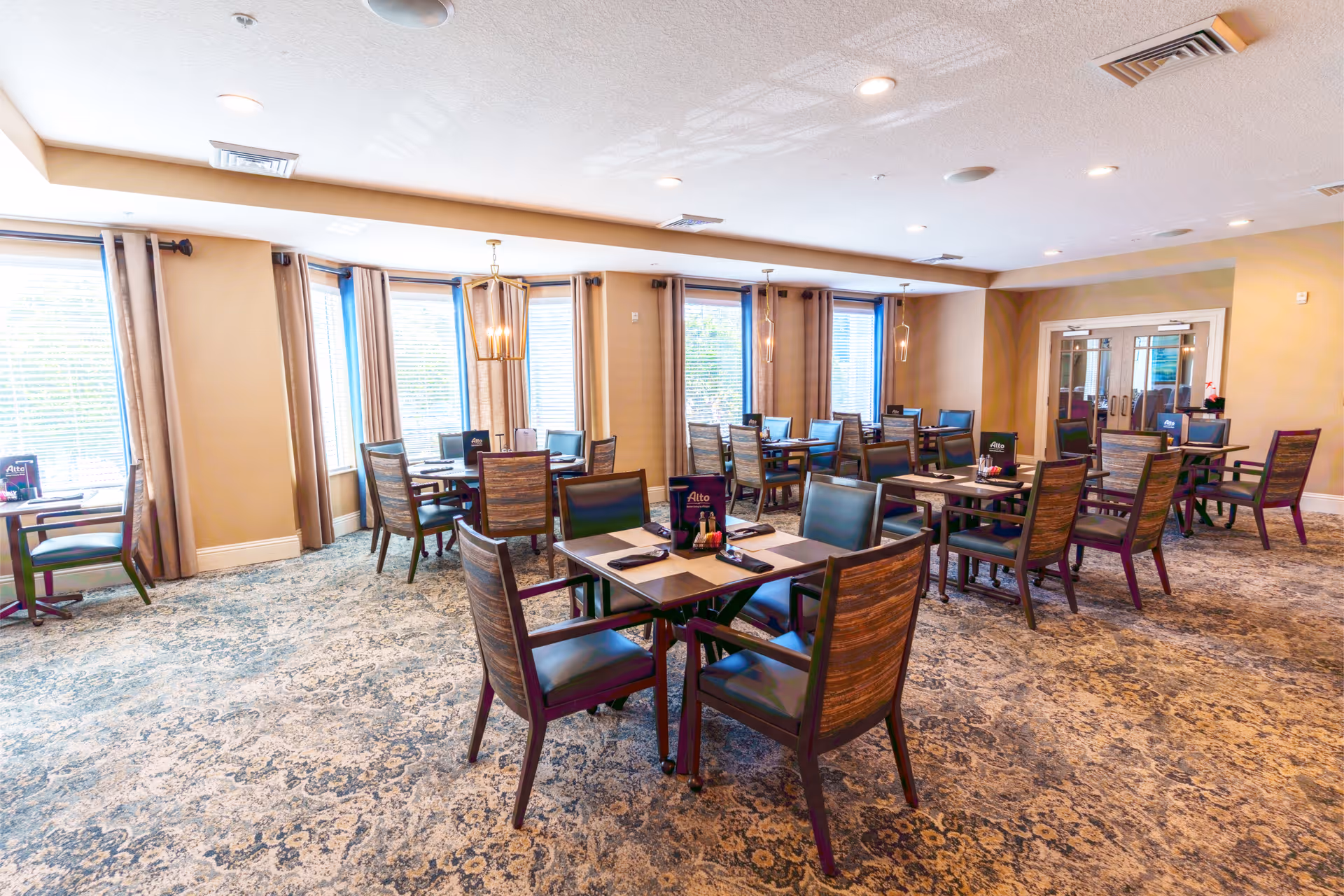 Bright dining room with multiple tables and chairs, large windows, and warm lighting.