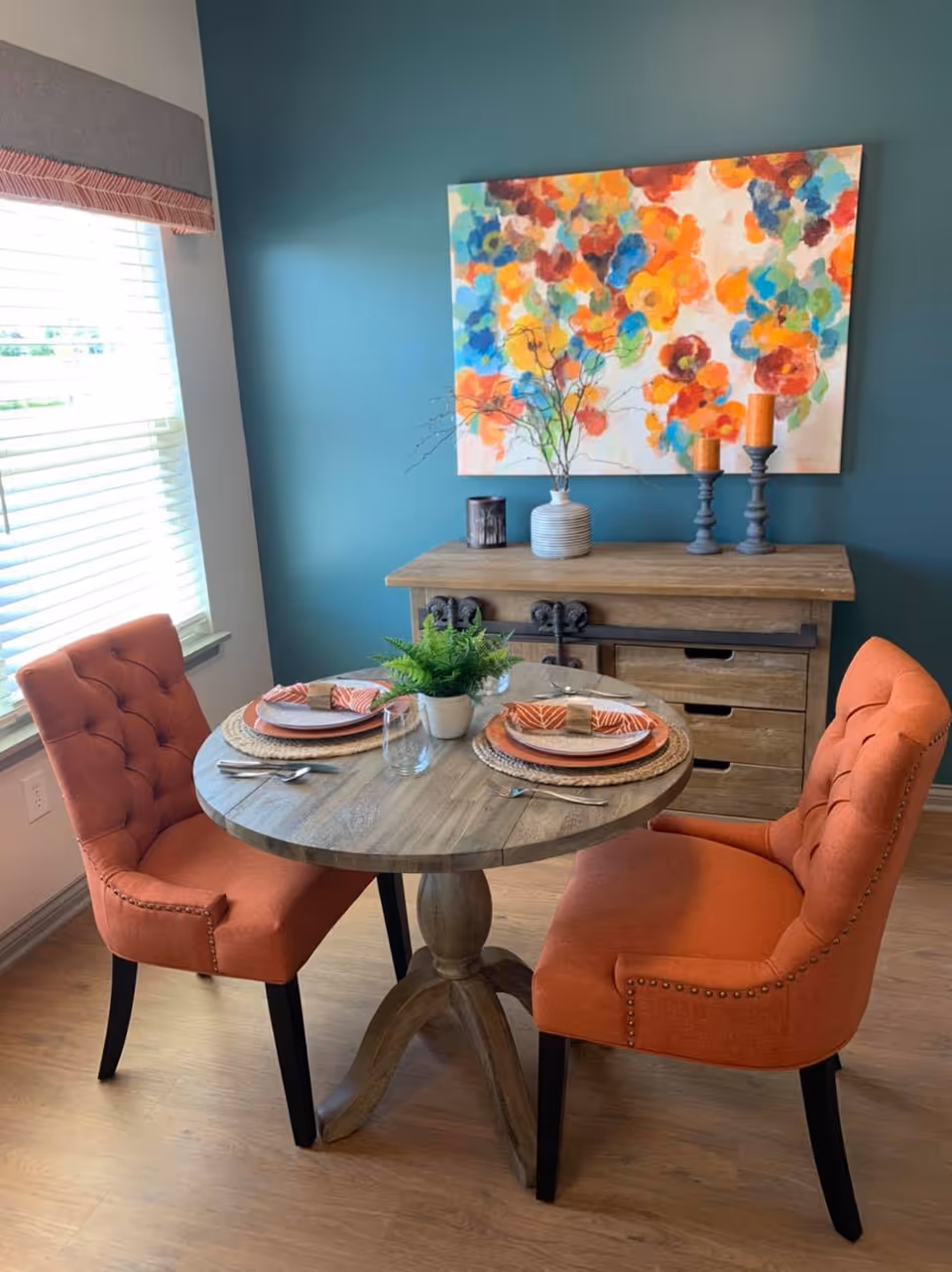 A small dining area with a round wooden table set for two, featuring orange cushioned chairs. The table is decorated with plates, napkins, cutlery, a glass, and a small potted plant. Behind the table is a wooden sideboard with decorative items including a vase with branches and two candle holders. A colorful abstract painting hangs on the teal-colored wall above the sideboard. A window with blinds and a valance is visible to the left.