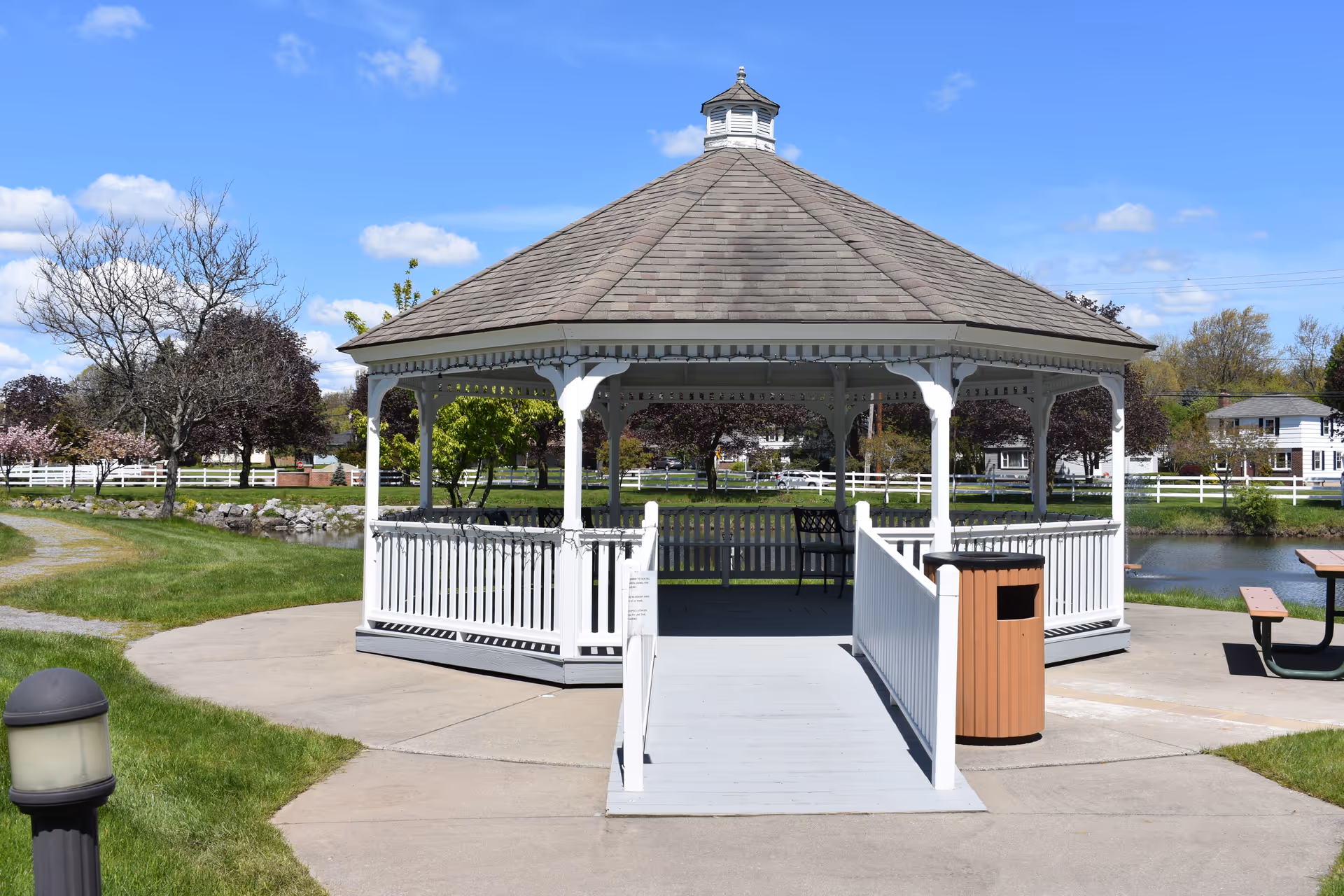 A white wooden gazebo with a shingled roof situated in a park-like outdoor area. The gazebo has a ramp leading up to it and is surrounded by green grass, trees, a pond, and a picnic table. The sky is clear and blue with a few clouds.