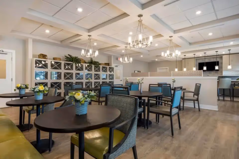 Bright, modern communal dining room with round tables and chairs, pendant chandeliers, a service counter and decorative shelving.