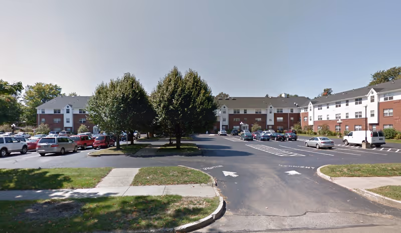 Exterior view of a senior living facility named Parkside Regency at South Shore with a large parking lot in front, several parked cars, and trees lining the driveway. The building is three stories tall with a combination of red brick and white siding.