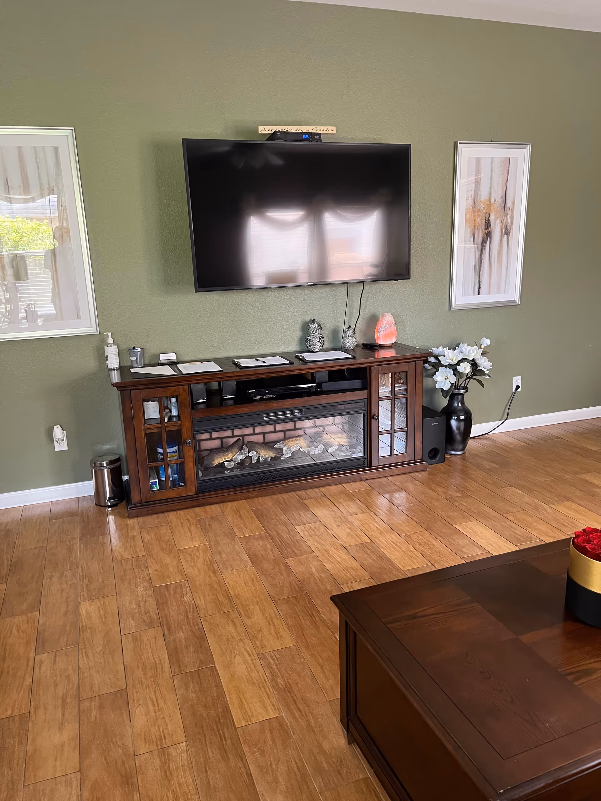A cozy living room area with a mounted flat-screen TV above a wooden entertainment center that includes an electric fireplace. The entertainment center has decorative items on top, including a salt lamp and two small sculptures. There are two framed abstract paintings on the green wall on either side of the TV. A black vase with white flowers is placed on the floor to the right of the entertainment center. The floor is wooden, and a wooden coffee table with a black and gold container holding red roses is partially visible in the foreground.